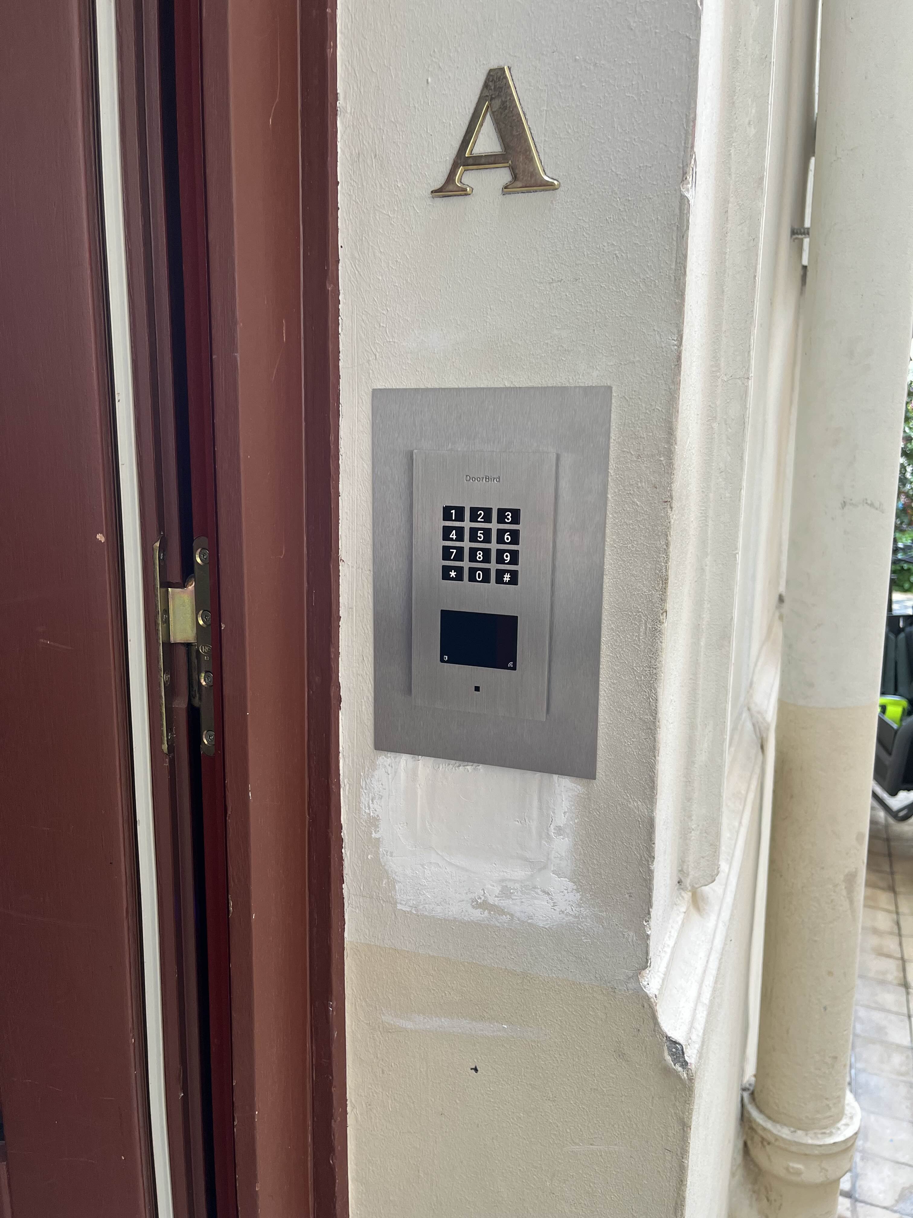 Metal DoorBird keypad mounted on a beige wall next to a brown door, with a gold letter A above it.