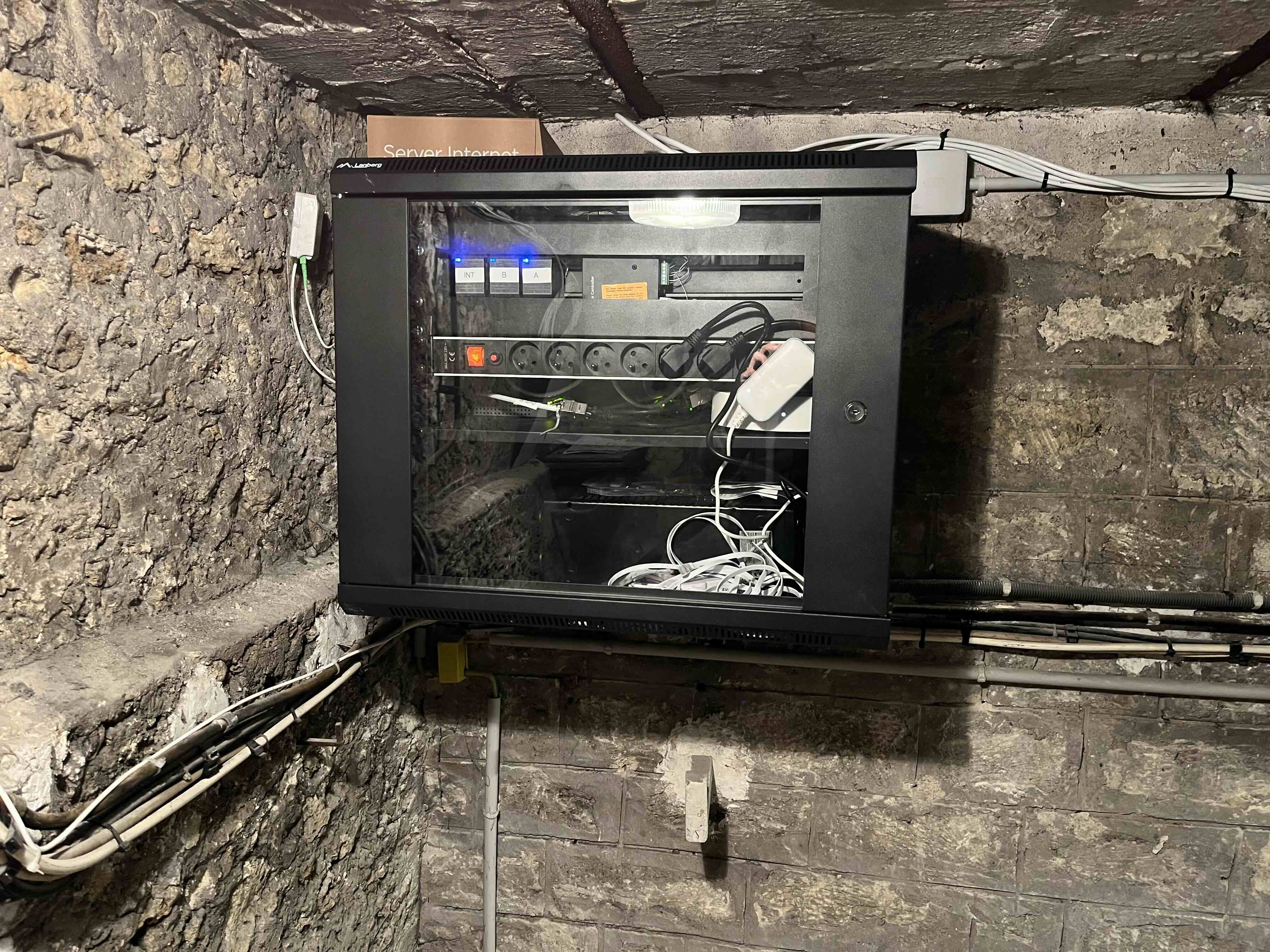 Wall-mounted black server cabinet with a glass door showing cables, power strip, and network devices inside on a stone wall background.