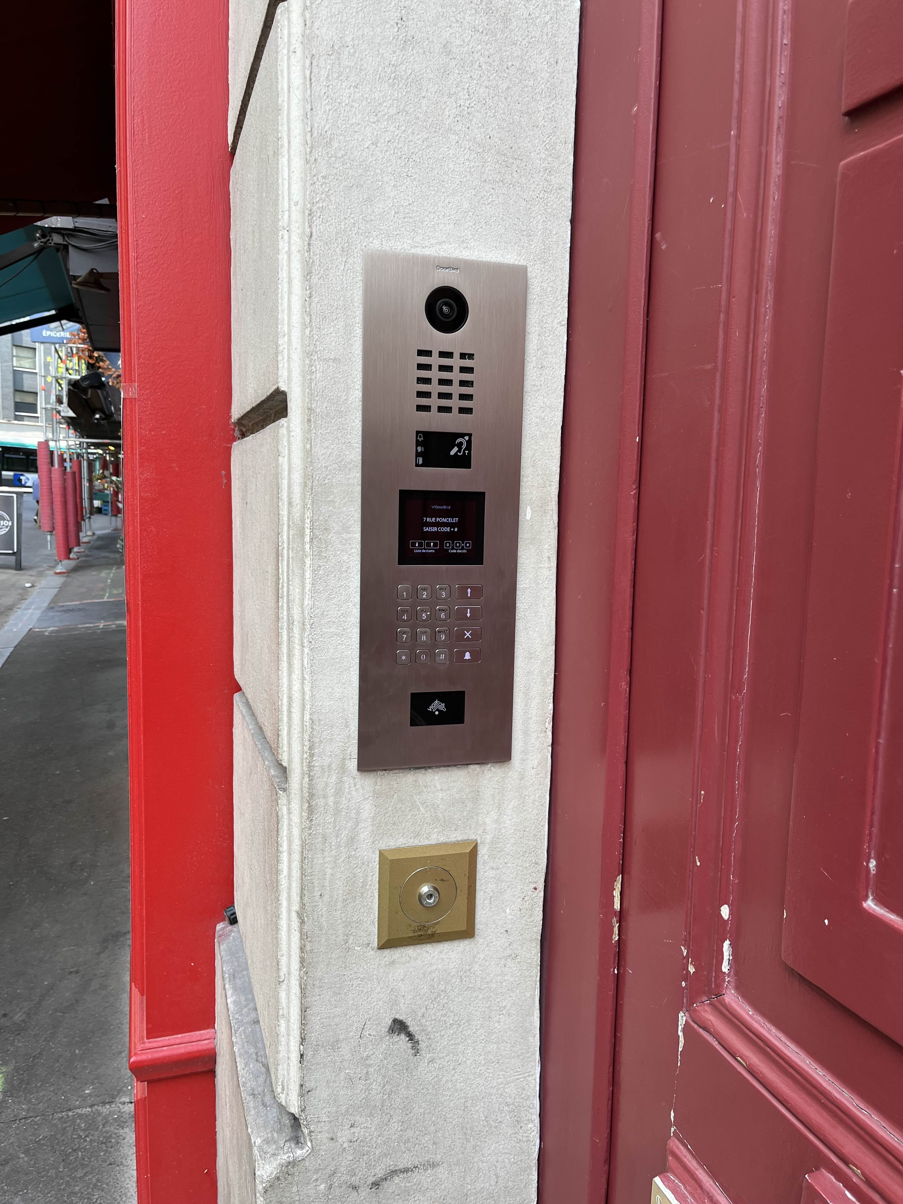 Modern video intercom keypad system mounted on a light-colored stone wall next to a red door frame in an urban sidewalk setting.