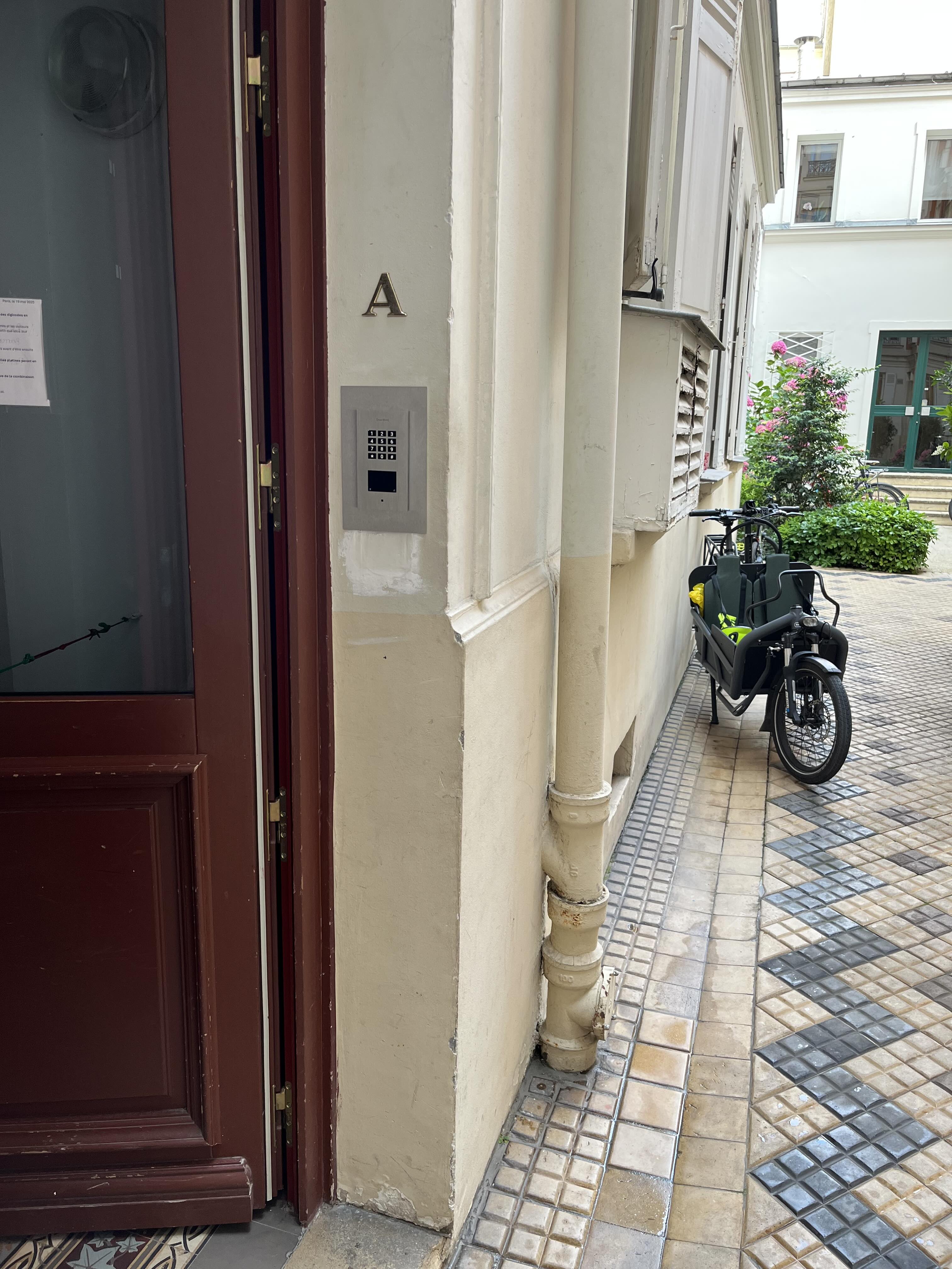 Entrance to building A with a keypad door lock, beige walls, tiled floor, and a cargo bike parked nearby.