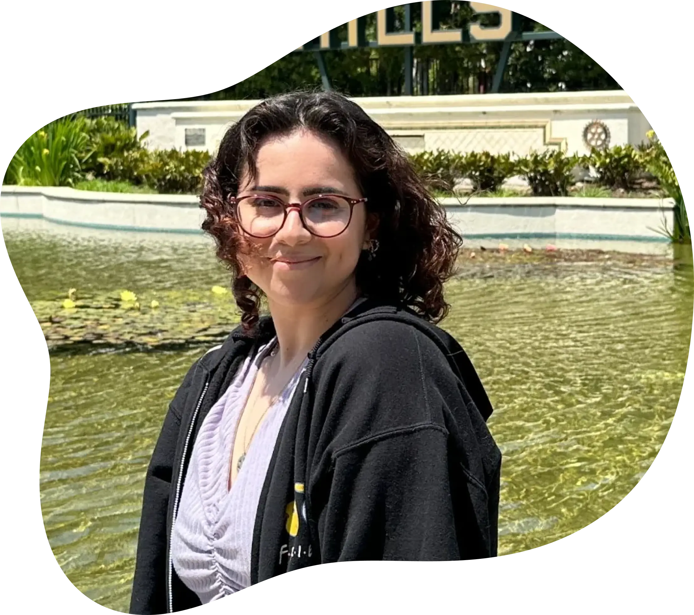 I am a woman with dark brown curly hair and glasses. I am pictured wearing a purple top and a black friends hoodie standing by a fountain smiling.