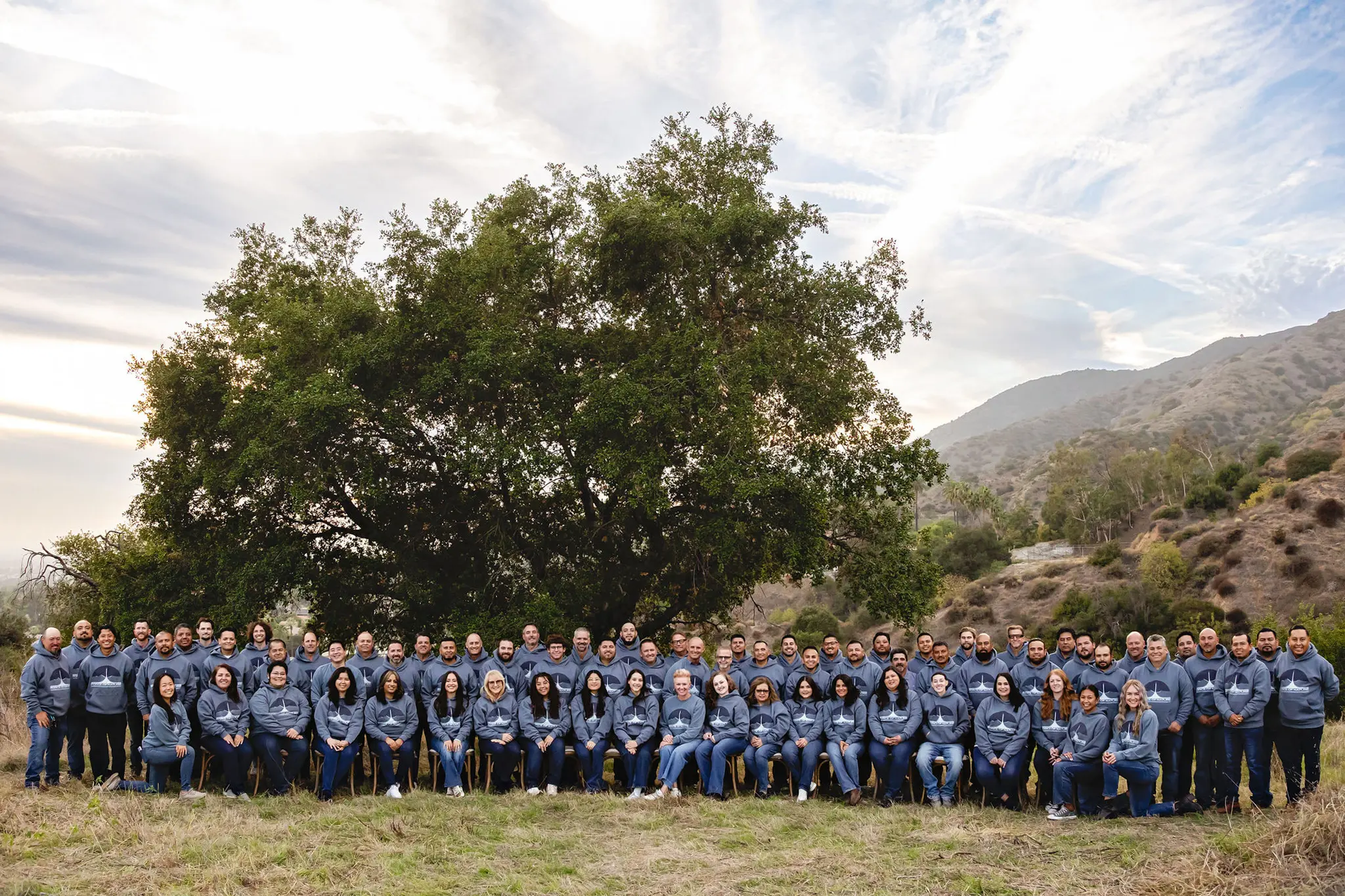 Large group of people outdoors wearing matching gray hoodies with a logo, posing in front of a large tree and hills.