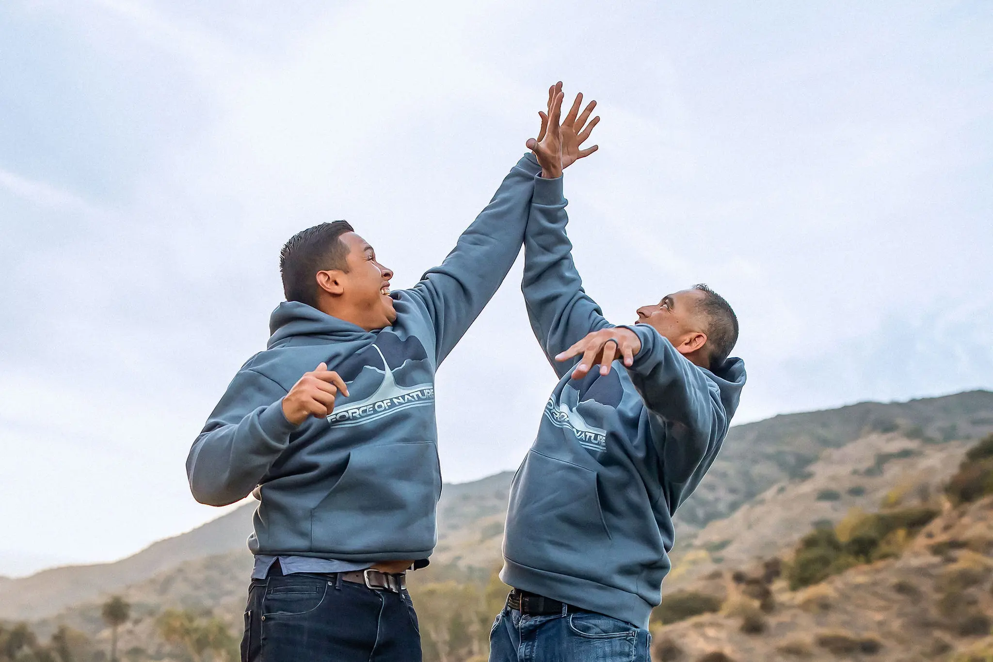 Two men wearing matching hoodies giving each other a high five outdoors with hills in the background.