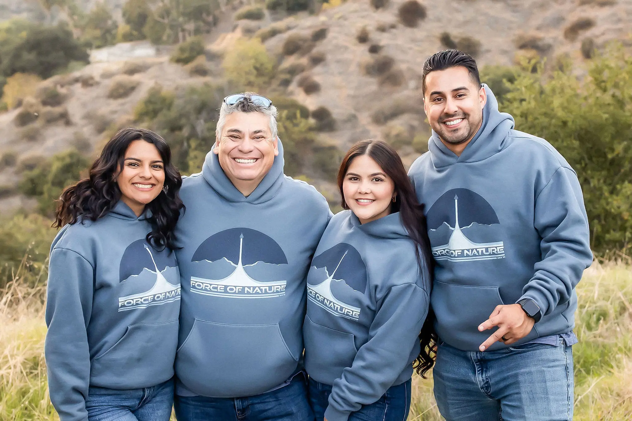 Four smiling people wearing matching blue hoodies with a 'Force of Nature' logo, standing outdoors with hills and greenery in the background.