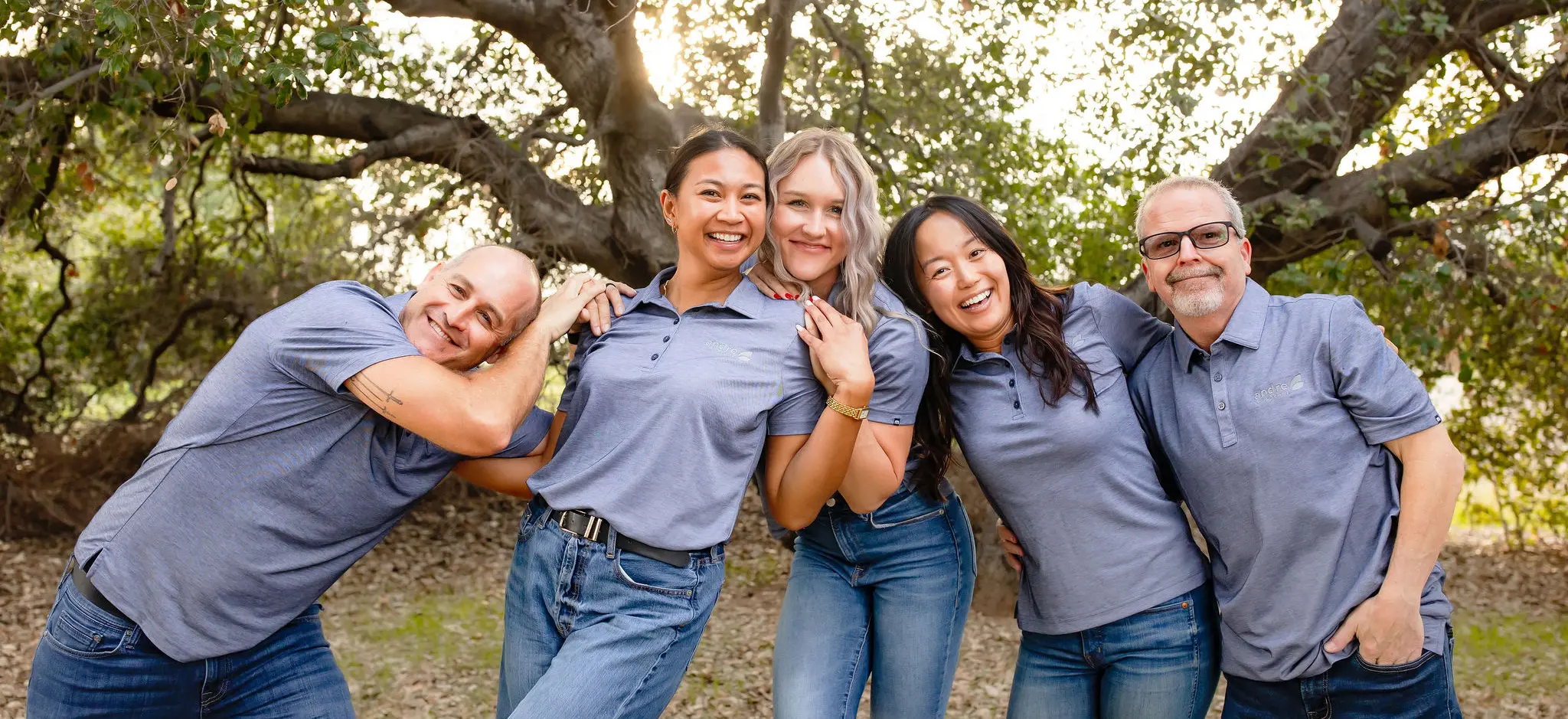 Five smiling people wearing matching light blue polo shirts and jeans standing closely together outdoors with trees in the background.