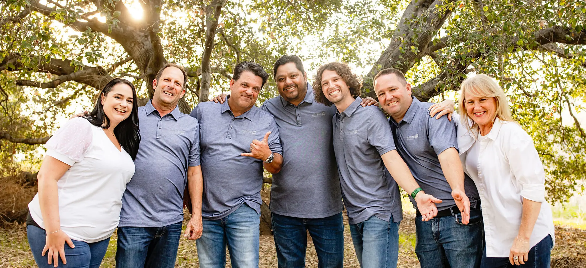 Seven adults smiling and posing together outdoors in casual blue and white shirts under leafy trees with sunlight filtering through.