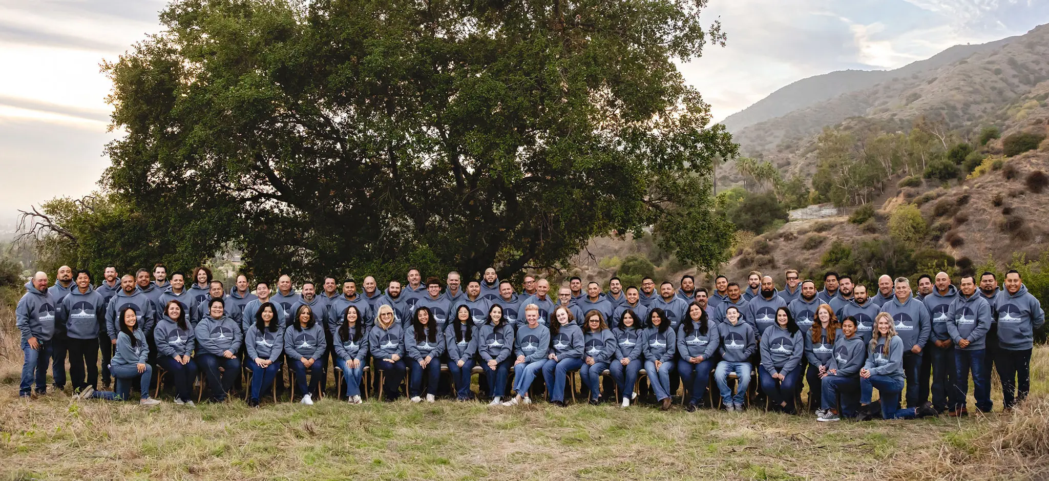 Large group of people outdoors wearing matching gray hoodies with a logo, posing in front of a large tree and hills.