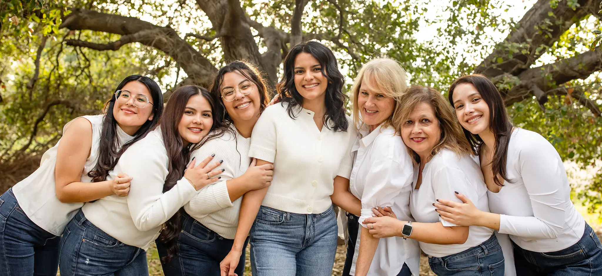 Group of seven women standing outdoors in front of trees, smiling and hugging each other.