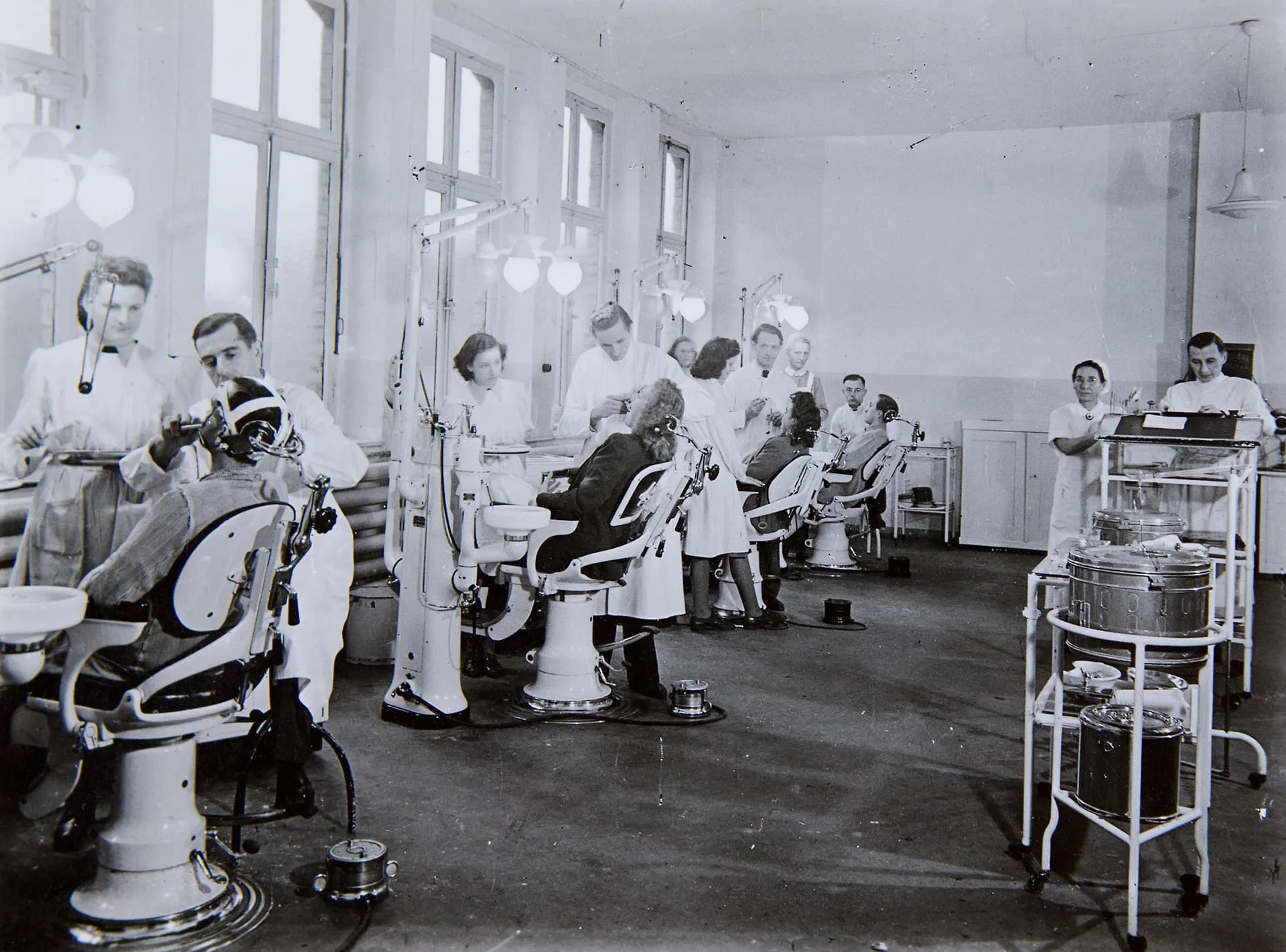 Historische Schwarz-Weiß-Aufnahme eines großen Behandlungsraums mit mehreren Zahnarztstühlen, Ärzten, Helferinnen und Patienten.