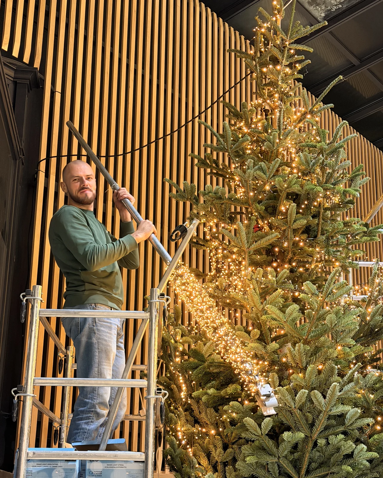 Un jardinier Captain Vegetal installe les guirlandes lumineuses d'un grand sapin de Noël depuis un échafaudage