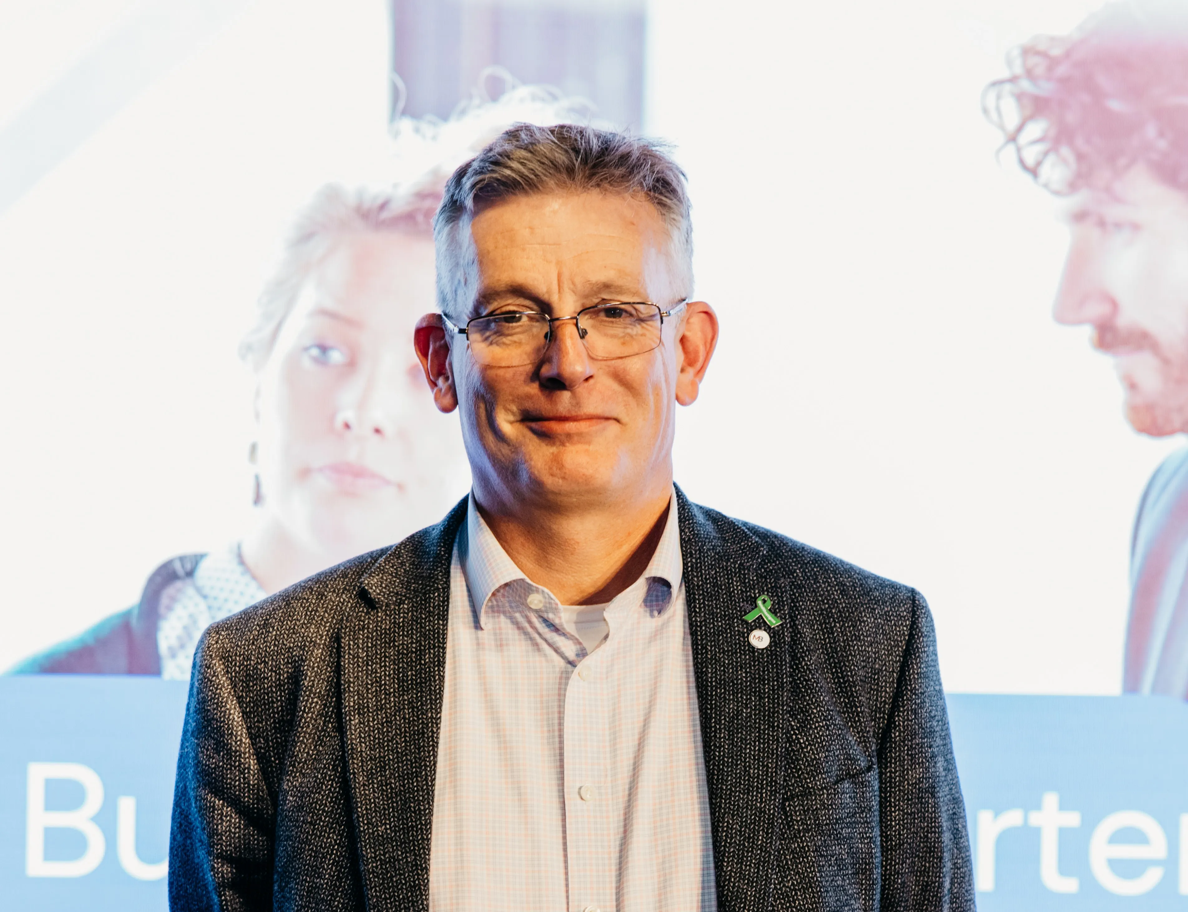 Middle-aged man with glasses and gray hair wearing a dark blazer and light shirt, smiling slightly in front of a blurred background with faces and text.