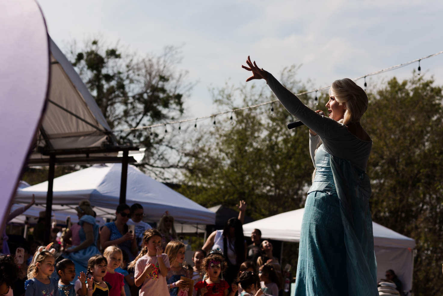 Performer dressed as Elsa engaging a crowd of children at an outdoor event with tents and string lights in the background.
