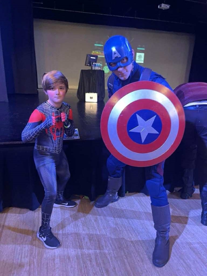 A child dressed as Spider-Man and a Captain America character performer posing together indoors on a stage floor.