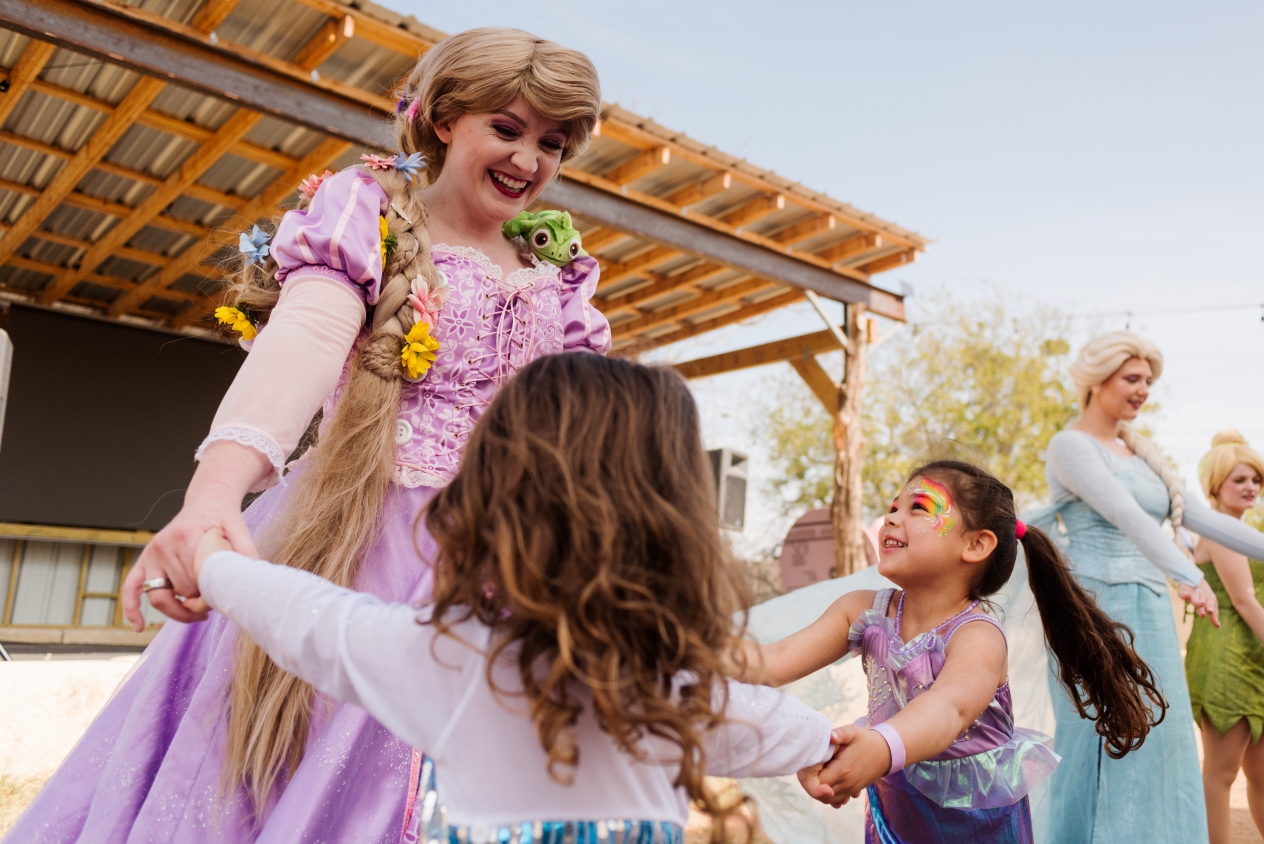 Woman dressed as Rapunzel holding hands and dancing with two young girls, with women dressed as Elsa and Tinker Bell in the background.
