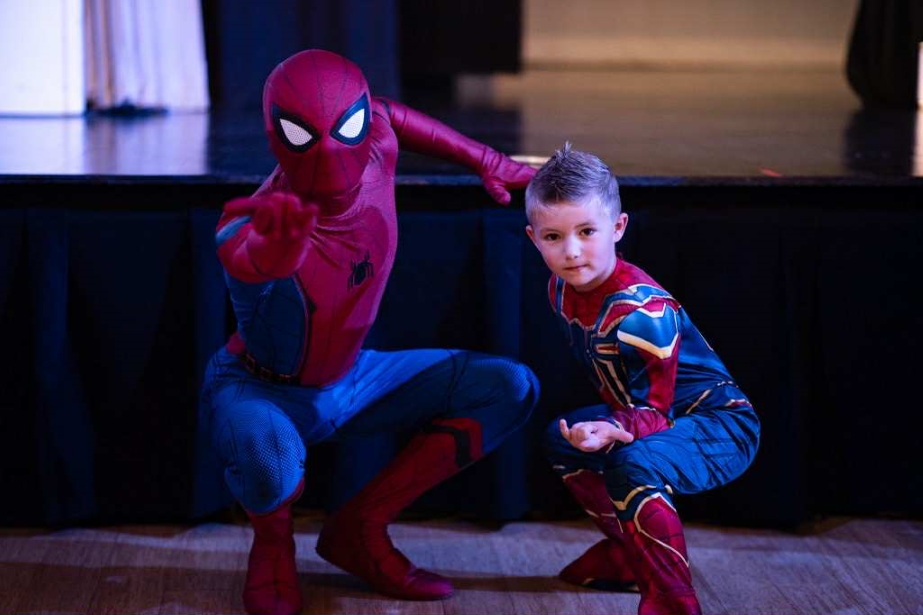 Child in Spider-Man costume next to Spider-Man performer, both crouching and posing indoors on a wooden floor.