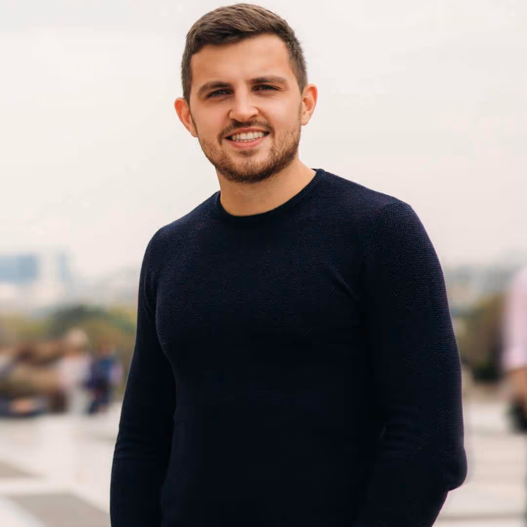 Jeune homme souriant portant un pull noir, debout à l'extérieur avec un arrière-plan flou urbain.