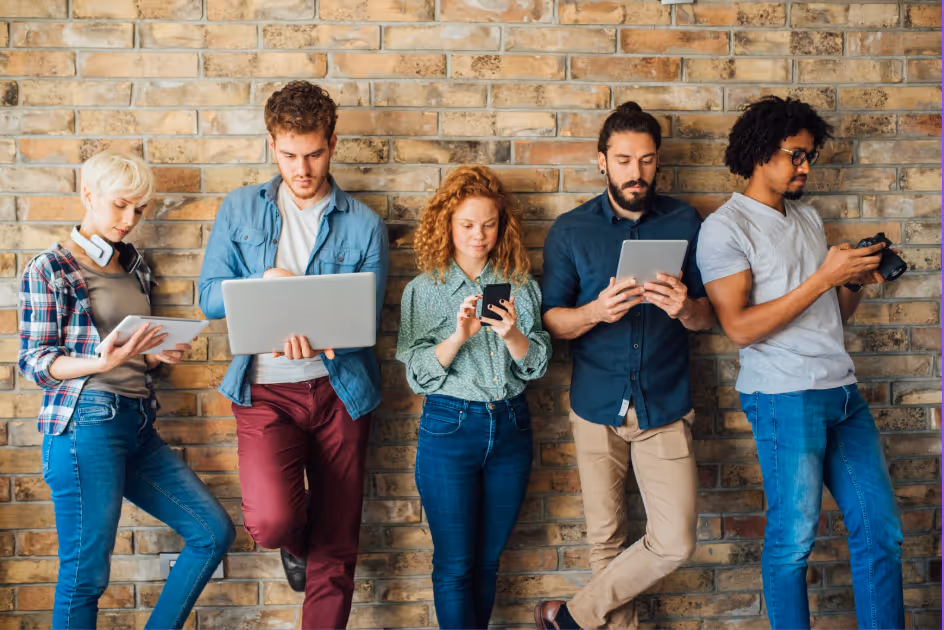 Eine diverse Gruppe junger Menschen steht vor einer Backsteinwand und nutzt verschiedene digitale Geräte wie Laptop, Tablet und Smartphone – symbolisch für modernes eLearning und Bewusstseinsbildung zu Unconscious Bias im Unternehmen.