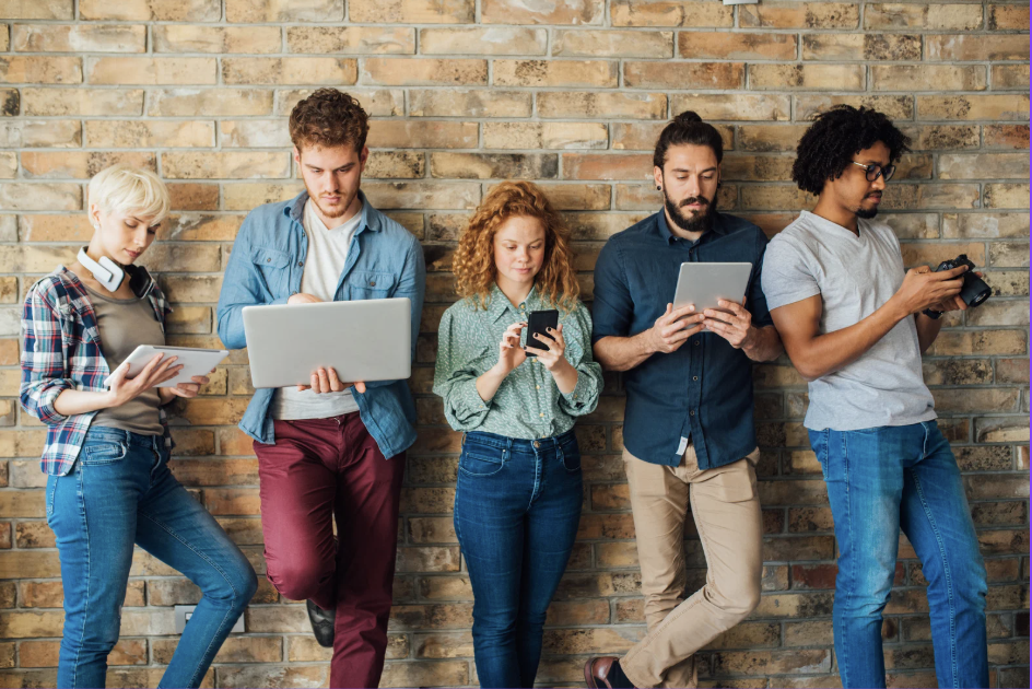 Eine diverse Gruppe junger Menschen steht vor einer Backsteinwand und nutzt verschiedene digitale Geräte wie Laptop, Tablet und Smartphone – symbolisch für modernes eLearning und Bewusstseinsbildung zu Unconscious Bias im Unternehmen.