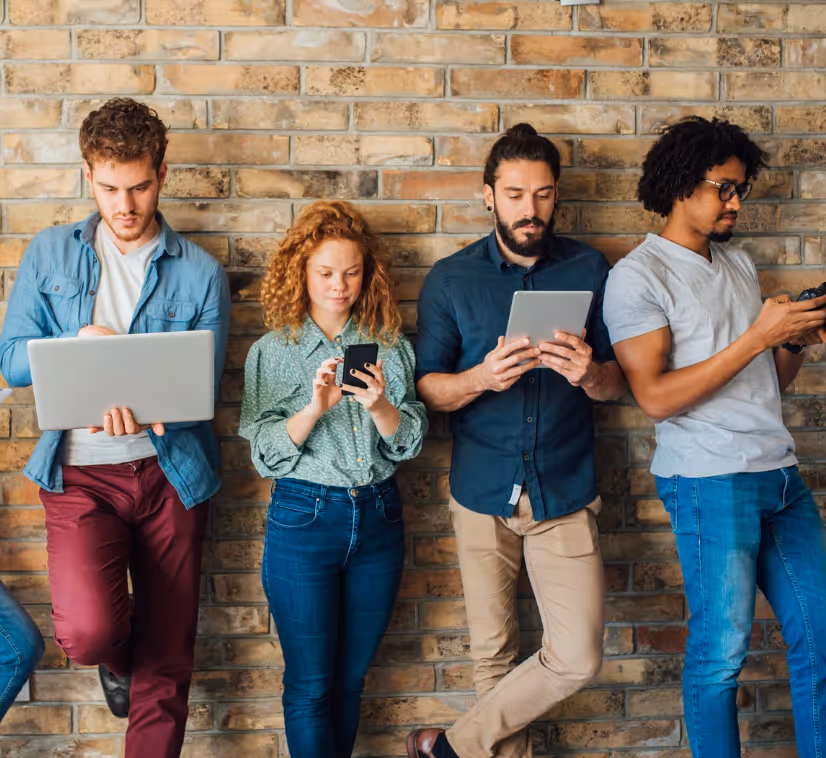 Eine diverse Gruppe junger Menschen steht vor einer Backsteinwand und nutzt verschiedene digitale Geräte wie Laptop, Tablet und Smartphone – symbolisch für modernes eLearning und Bewusstseinsbildung zu Unconscious Bias im Unternehmen.