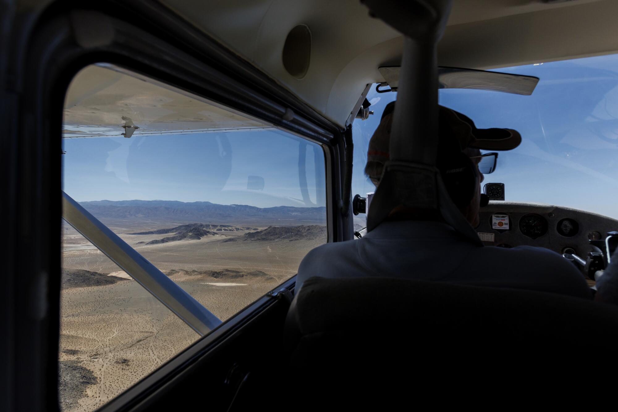 A pilot is silhouetted by blue sky as they fly a small airplane over the desert.