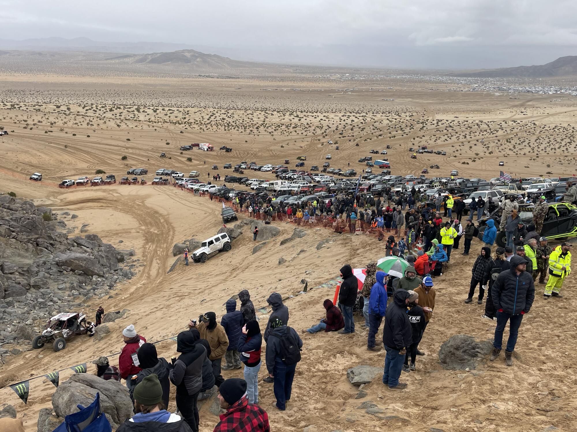 Spectators on a desert ridge watch a motor vehicle contest.