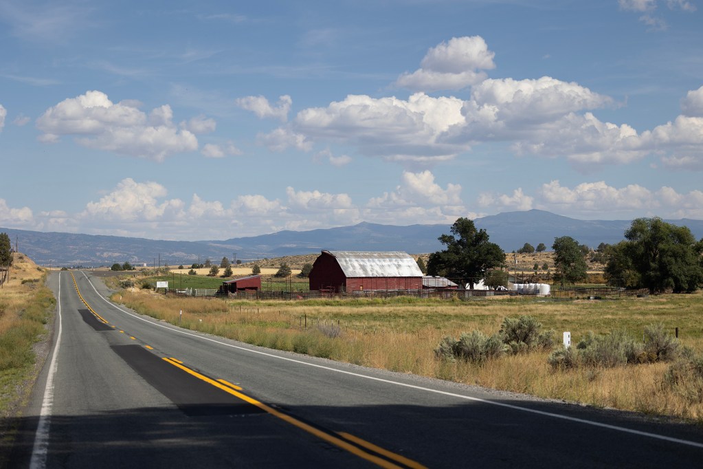 A long two-lane road stretches into the distance under a partly cloudy blue sky. To the right of the road sits a red barn with a silver metal roof, surrounded by fencing and open fields. Rolling hills and distant mountains line the horizon.