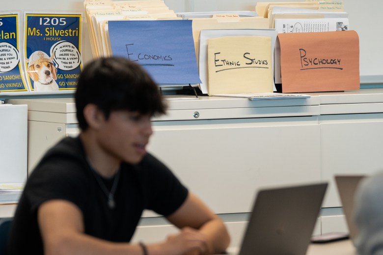 Manila envelopes that are labeled "economics," "ethnic studies" and "psychology" are displayed on top of a cabinet inside a high school classroom. A student can be seen int he foreground using their laptop.