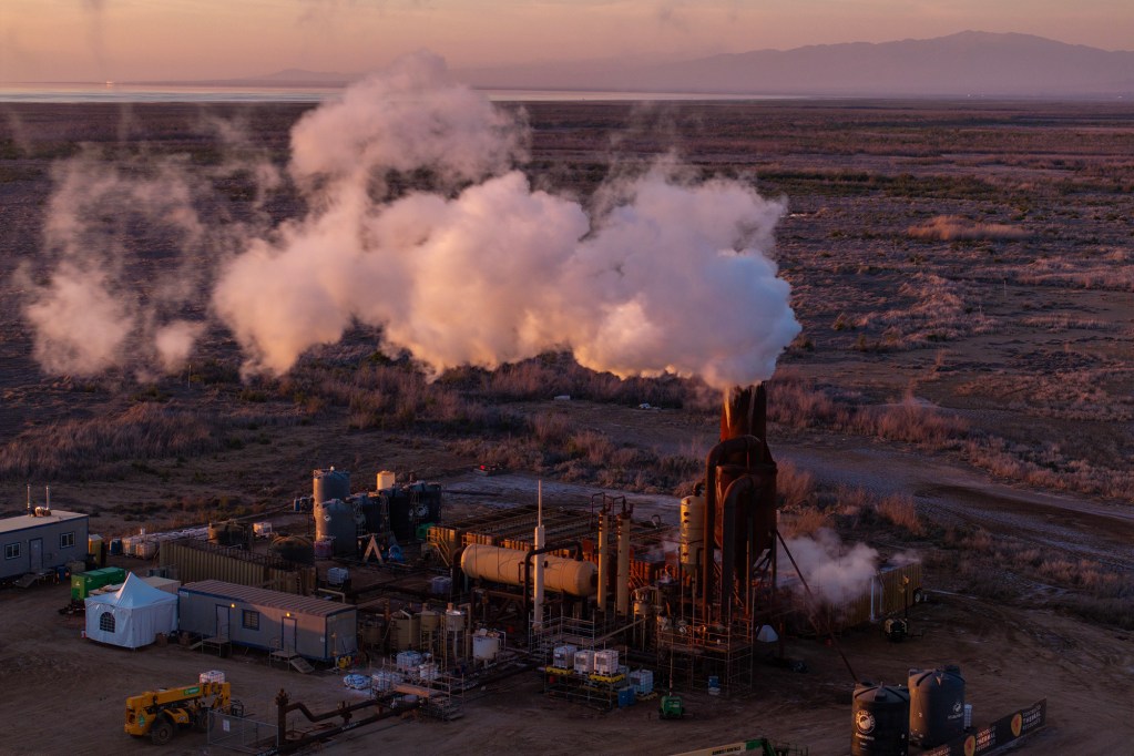 An industrial geothermal plant emits thick white steam into the air at sunset, with a vast, arid landscape stretching into the distance. The facility consists of metal pipes, large storage tanks, and temporary structures, with scattered equipment on the dirt ground. A mountain range and a body of water are visible on the horizon, bathed in soft golden light.
