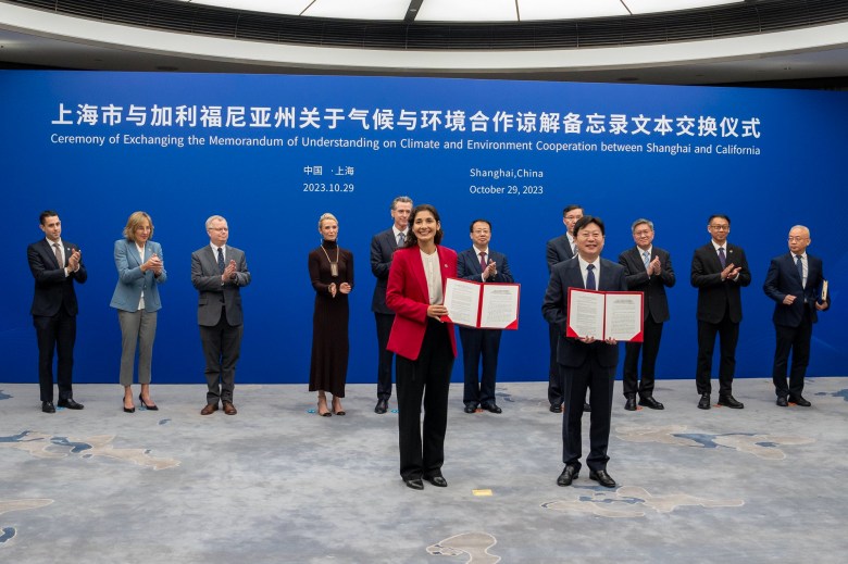 A group of people clap while standing in front of a large blue wall with Mandarin and English writing that reads: "Ceremony of Exchanging the Memorandum of Understanding on climate and Enviroment Cooperation between Shanghai and California" Two people, one wearing a red blazer and another wearing a black suit, stand in front of the rest.