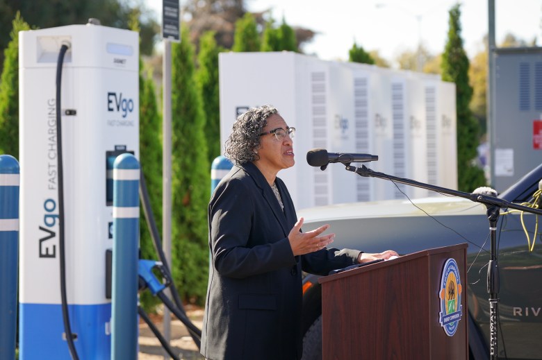 A side-view of a person, with gray curly hair and wearing a black blazer, as they stand in front of a podium next to several electric vehicle chargers during a press conference.