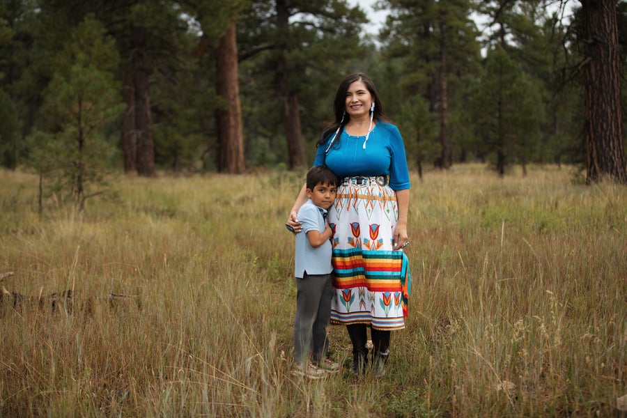 Native American mother wearing a colorful ribbon skirt, standing beside her young son with a forest in the background