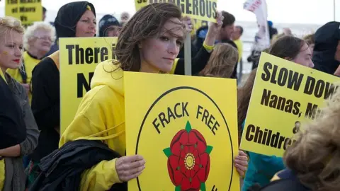 Getty Images A woman with brown hair and wearing a yellow jumper holds a yellow sign with the Lancashire rose in the centre and "frack free" written in black writing above it. Around her are other protestors with similar signs.