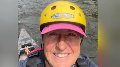 Justine Evans Wildlife filmmaker Justine Evans takes a selfie while canoeing on the river Wye. She is sat in a black kayak, wearing a dark blue hoodie, a red life jacket and a pink baseball cap.