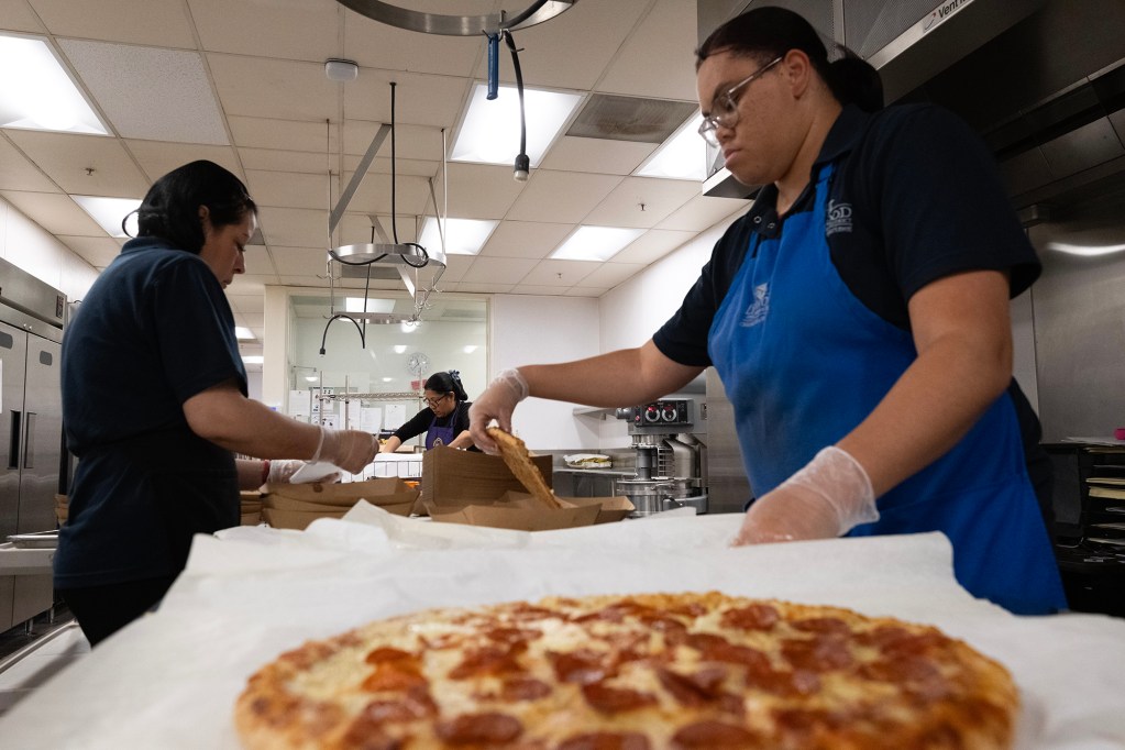 Three cafeteria workers in uniforms and gloves prepare pepperoni pizza slices in a commercial kitchen. One worker in the foreground places a slice into a cardboard container, while others pack meals in the background under bright fluorescent lighting.