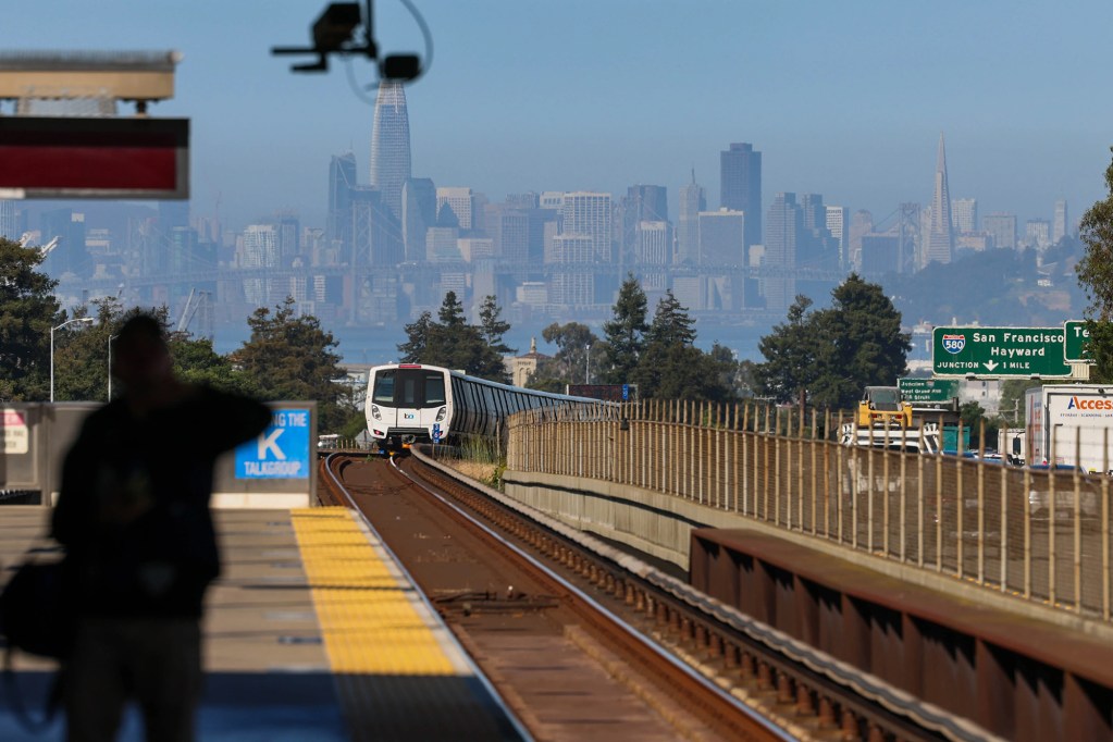 A white train in makes it's way down train tracks towards a station with a person waiting. The San Francisco skyline can be seen in the background.