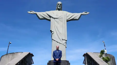 PA Media Prince William stood in front of the Christ the Redeemer statue, which looks light grey. The sky is bright blue and the prince is wearing a navy suit.