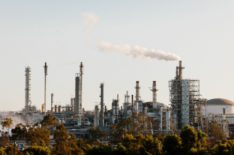 A wide view of a refinery location on a clear day with various structures, including distillation columns, as smoke is released into the air.
