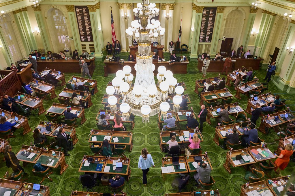 A wide-view from above of a legislative room filled with lawmakers sitting at their desks. A large crystal chandelier hangs from the ceiling. The room is decorated with beige walls, mint-color columns, gold fixtures and a lime green carpet.
