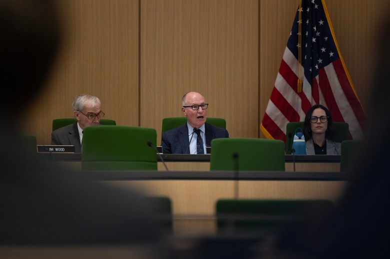 A panel of legislators sits at a long dais with green chairs inside a hearing room. An American flag stands behind them. A person at the center is speaking into a microphone while wearing a suit and glasses.