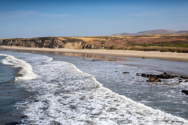 Aerial view of a stretch of coastline with waves moving towards a sandy beach line, spread throughout the frame, and with a bluff of cliffs just beyond the sand.