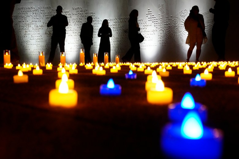 Silhouettes of people standing around a lit-up memorial inside a museum with rows of plastic candles lit up on the ground.