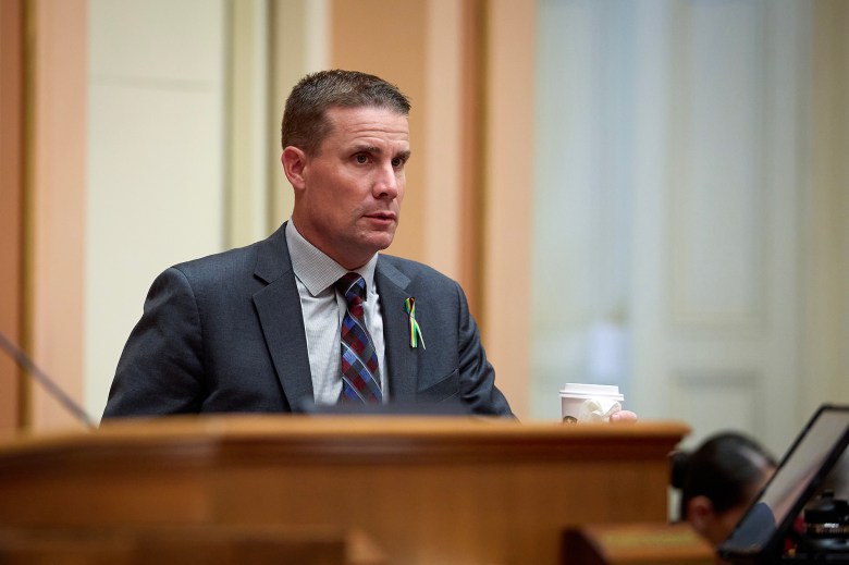 A person in a suit and plaid tie stands at a podium inside a government building, holding a cup of coffee. A multicolored ribbon is pinned to their lapel. The setting appears formal, with soft lighting and wood paneling in the background.