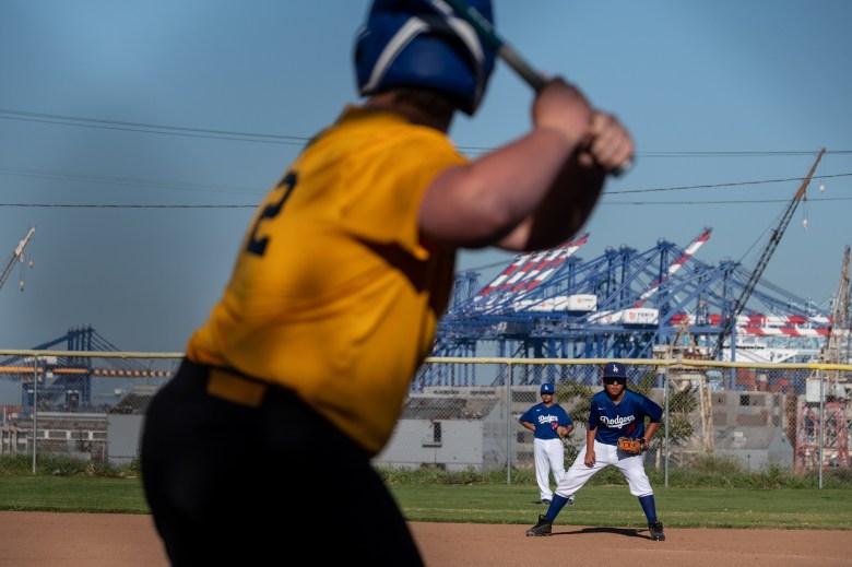 Two young baseball players wearing blue and white jerseys stand in their positions as they watch a batter get ready for a pitch during a game. Blue cranes from a nearby port can be seen in the distant background.