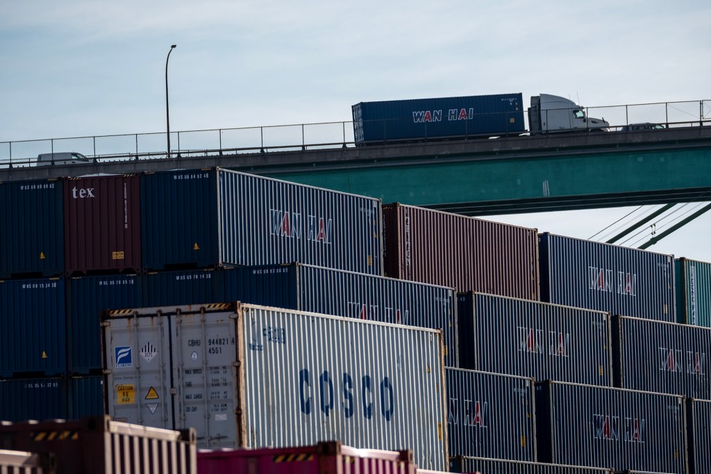A stack of multicolored shipping containers, including brands like COSCO, Wan Hai, and tex, sits at the Port of Los Angeles. Above, a semi-truck carrying a blue container drives across a bridge, silhouetted against a light sky. The composition emphasizes the scale and flow of international freight transport.
