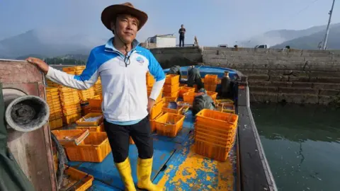 BBC/Hosu Lee A man wearing a hat, a light jacket and bright yellow boots stands on the deck of a moored boat, alongside lots of yellow crates.