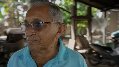 BBC / Tony Jolliffe A man with siver hair and tanned skin wearing dark sunglasses and a light blue polo neck t-shirt stands in his farmyeard. In the background, you can see his wooden tools and a motorbike. 