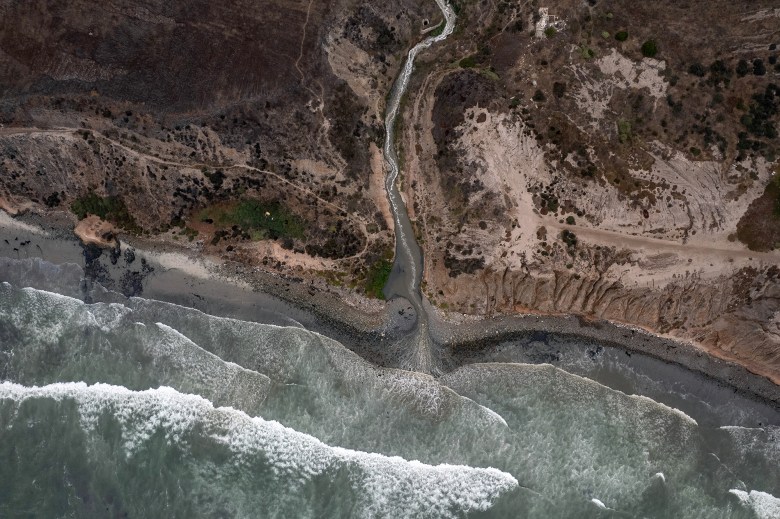 An aerial view showing a wastewater river, surrounded by dirt terrain, that spills into an ocean as waves crash onto the shore.