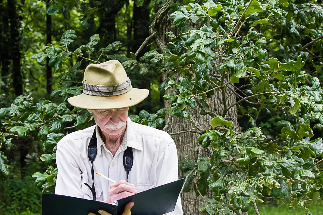 a man with a white shirt and brimmed hat looks at a binder in front of greenery