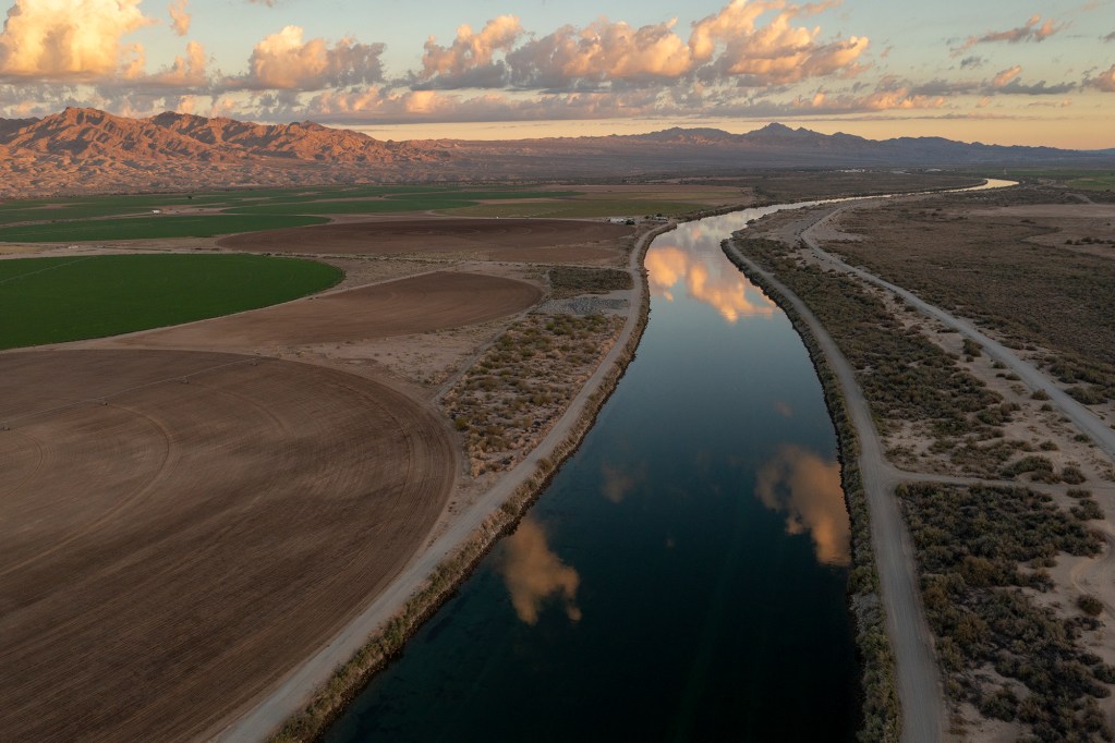 An aerial view of the Colorado River as it winds through grass fields on farmland with clouds reflecting in the river from the sky. A mountain range is shown in the background, with rays of light hitting it at sunset.