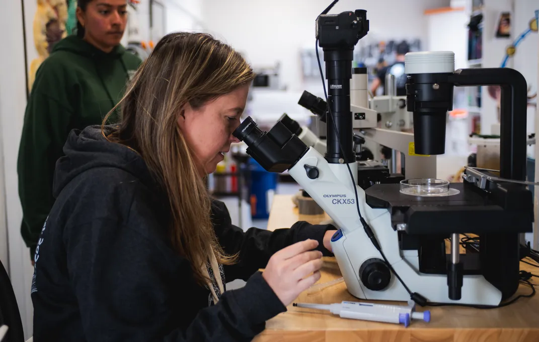 a woman sits at a table looking through a microscope